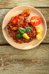 Chicken legs with red rice in tomato-garlic sauce on a plate on a wooden table. Copy space.