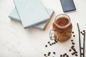 Mason jar of tasty iced coffee with notebooks and mobile phone on table