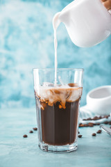 Pouring of milk into cold coffee in glass on color background