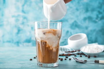 Pouring of milk into cold coffee in glass on color background