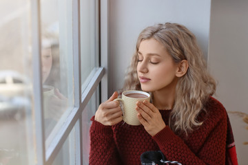 Beautiful young woman drinking hot chocolate near window at home