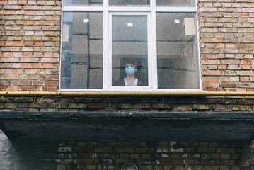 a guy in a protective mask stands in a brick house outside the window and looks outside. Stay home, concept, coronavirus 2020.