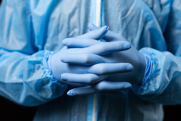 crossed fingers of male hands in medical disposable gloves and chemical protective overalls, blue, close-up on a dark background