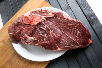Fototapeta premium A piece of fresh meat on a white plate. Beef steak. Meat cutting. A wooden cutting Board, on a dark table.