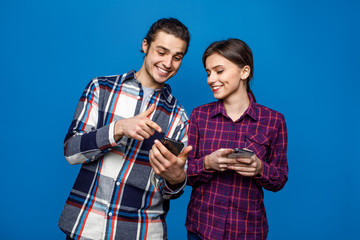 Happy beautiful couple wearing  t-shirts standing isolated over blue background, playing games on mobile phone