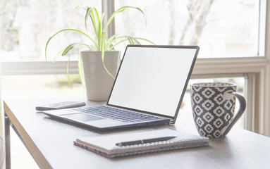 Minimalist home office set up, desk with a laptop with copy space, green plant, cell phone, notebook a pen and coffee mug against a bright window with natural light