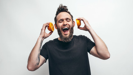 Young bearded man holding orange slices in hands in front of his ears like headphones for listening...