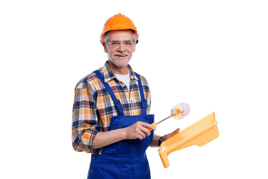 Portrait of senior male builder being going to paint walls in house, holding paint roller in hand, wearing special uniform, apron and protective helmet, isolated on white background