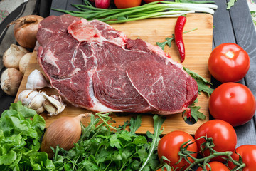 The preparation of fresh meat. Fresh beef steak on a wooden table. Meat surrounded by spices and vegetables. Fresh fruit. Red ripe tomatoes. Dietary food.