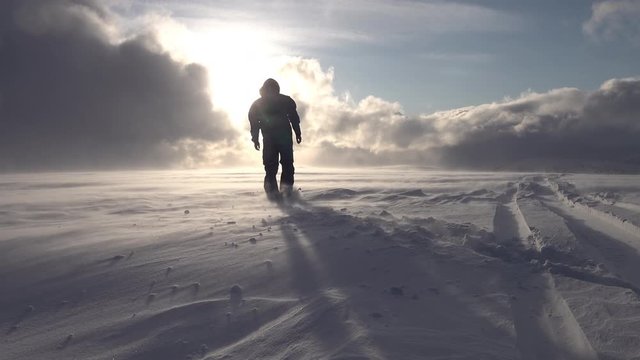 Silhouette of man in black snowmobile overall and helmet, he walking towards Sun shining though heavy clouds in mountains, poor snow visibility, high speed snow drift conditions