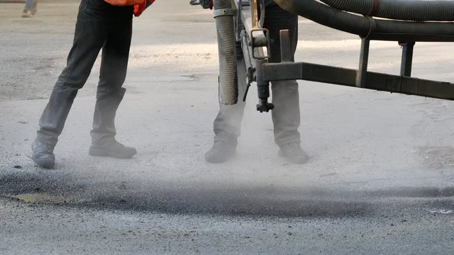 Workers perform minor repairs to pavement by spraying asphalt chips into pits on roadway. Machine blows hot bitumen mixture as road patches. Sealing cracks by blowing crushed granite stone with tar 