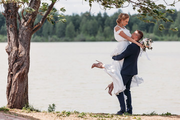 Panoramic view at young couple bride in white and groom in blue standing among lake under a branch of green tree, posing and embracing. The groom lifted bride in his arms