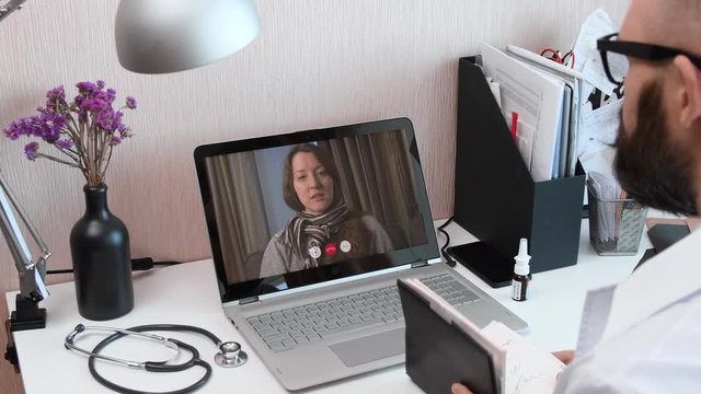 Telemedicine. Consultation with physician through mobile video call. Doctor talking to comlaining patient using video chat application on laptop and writes down symptoms.