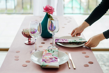 Close up woman's hands serving party table in gentle pink colors with white dishes, glasses for wine, floral textile napkin. Happy birthday or baby shower for girl.