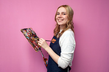 girl artist holds brushes and a palette and smiles on a pink background, student of art school,...