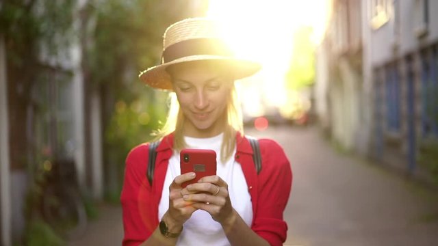 Selective focus on smartphone. Positive smiling hipster girl in hat feeling happy of received message with good news, cheerful millennial female enjoying chatting online via app while standing outdoor