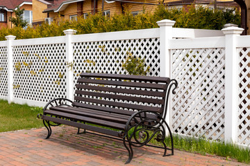 Empty dark wooden bench on the background of a white plastic fence in a rural village. Comfortable resting place under the open sky. Close-up, copy space.