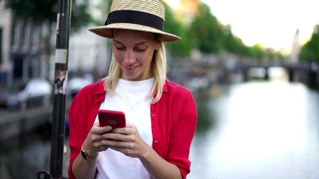 Happy smiling hipster girl in stylish hat enjoying communication while using mobile phone, positive female tourist text messaging via application on smartphone while standing outdoors on city street
