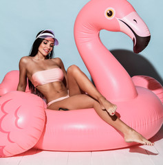 Young tanned slim brunette woman in sexy pink beach bikini and cap sitting on big pink flamingo and...