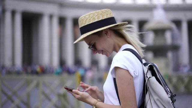 Cheerful woman with backpack taking picture during walking on city square with exciting buildings using smartphone camera with high quality, positive hipster girl chatting online via application 
