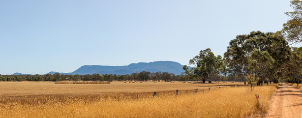 Panoramic view of rolling farm fields of freshly harvested hay on rural properties in Victoria with part of the Grampians National Park mountain range rising in the background