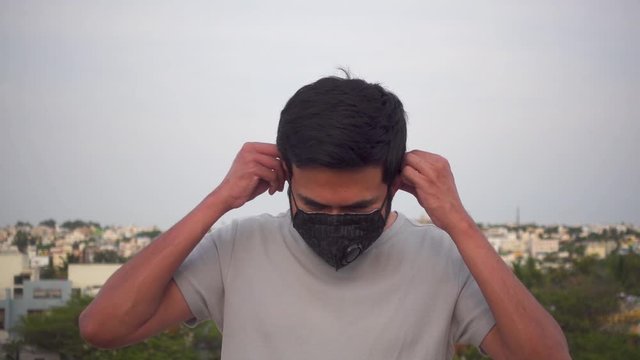 Mid shot of a smiling young Indian man in a tee shirt wearing a black pollution mask and showing a thumbs up
