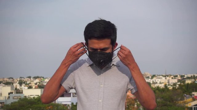 Mid shot of a serious young Indian man in a casual shirt wearing a black pollution mask and turning his head