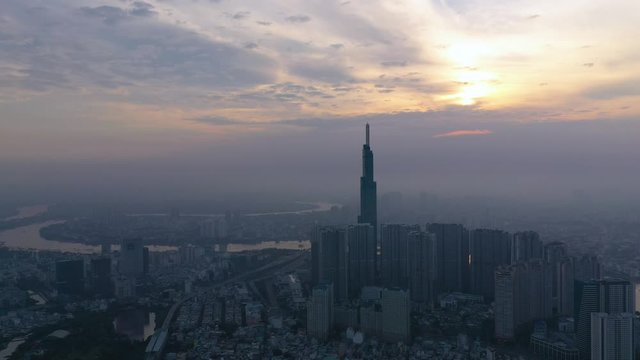 Sunrise with smoggy morning panoramic drone shot over densely populated urban area of a modern city with high rise towers in silhouette. Camera flies in slowly