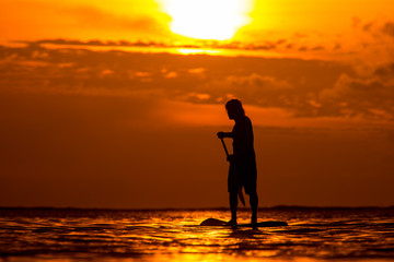 surfer rides by paddle board (S.U.P.) in the ocean against the background of a large disk of the setting sun