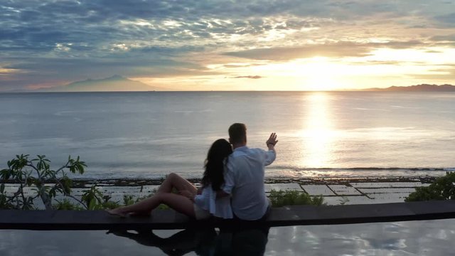 Couple In Love At Luxury Resort On Romantic Summer Vacation. People Relaxing Together In Edge Swimming Pool , Enjoying Beautiful sunset Sea View. Happy Lovers On Honeymoon Travel. Relationship, Romanc