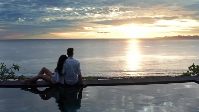 Couple In Love At Luxury Resort On Romantic Summer Vacation. People Relaxing Together In Edge Swimming Pool , Enjoying Beautiful sunset Sea View. Happy Lovers On Honeymoon Travel. Relationship, Romanc