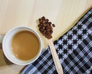 A white coffee mug with coffee in it and a spoonful of roasted coffee beans laid on a wooden...