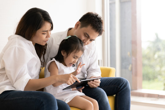 Happy Asian Family Using Technologies For Fun. Father, Mother And Daughter Looking At Digital Tablet On Sofa In The Home. Communication Technology And Lifestyle