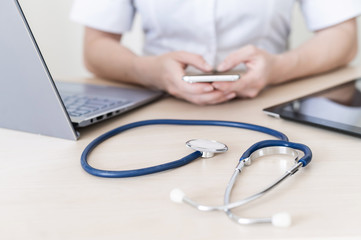 Unrecognizable female doctor sits at a desk and uses a smartphone. Closeup of the hands of a practitioner with a phone. Stethoscope tablet and laptop on the table.
