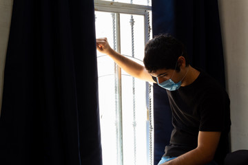 Fototapeta na wymiar young man wearing a blue medical mask looking out of a window very sad about the coronavirus quarantine