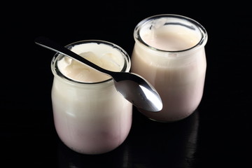 Yogurt in open small glass jar with spoon at black background.