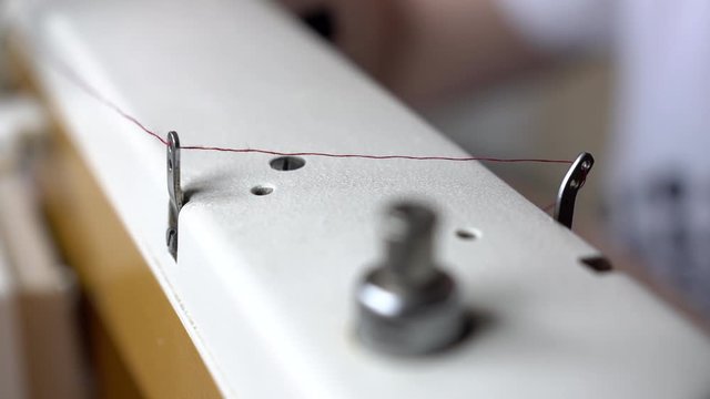 Thin red thread transporting over white sewing machine down to the needle with worker sewing in the background. Thin red line. Process of sewing on the sewing machine