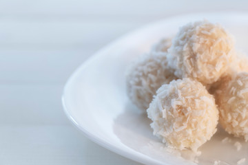 Coconut candy balls in plate covered with chocolate and shredded coconut on rustic wooden background.