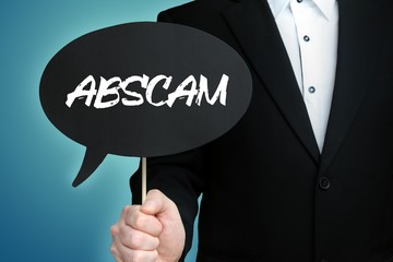 ABSCAM. Lawyer in suit holds speech bubble at camera. The term ABSCAM is in the sign. Symbol for law, justice, judgement