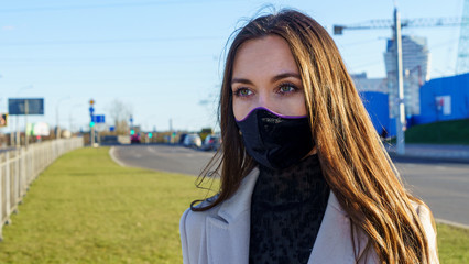 Coronavirus fashionable medical face mask worn by young female with dark long hair, standing at tram stop on a urban city street on dusk. Stop covid 19 pandemic or air pollution concept.