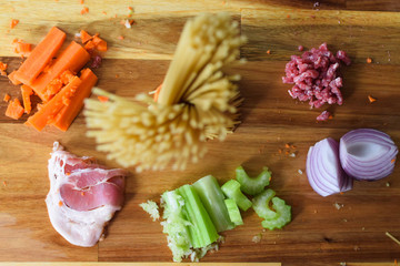 Spaghetti Bolognese Pasta ingredients which include uncooked pasta, carrots, ground pork, green celery, bacon, and garlic on Wooden Background. Selective focus with raw pasta at the center