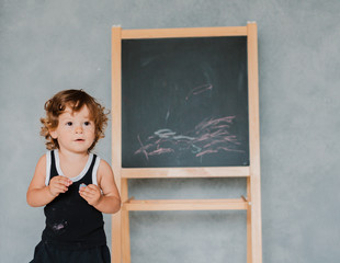 small child draws with chalk on a black chalk Board at home in the nursery against a gray wall.