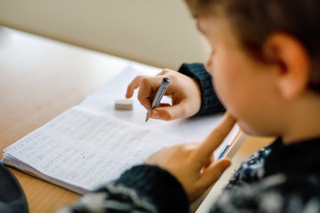 Hard-working happy school kid boy making homework during quarantine time from corona pandemic disease. Healthy child writing with pen, staying at home. Homeschooling concept