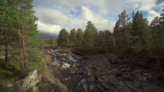 A shallow mountain river in the Trollheimen park carrying its waters gently through the rocky canyon down the valley.  Pine trees stand on the river banks. Thick white clouds covering the sky.