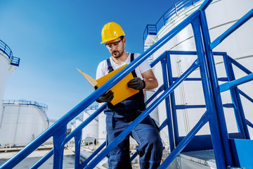 Hardworking handsome caucasian worker in overall and with helmet on head standing on the stairs and...