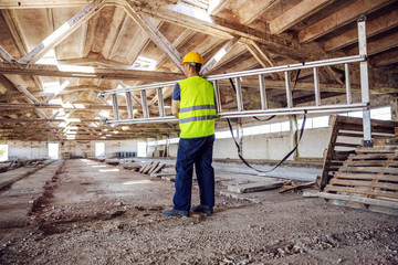 Rear view of construction site worker relocating ladder while walking on site.
