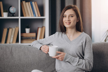 Fototapeta premium Portrait of happy woman in casual clothing sitting on grey couch with cup of coffee and looking at camera. Charming girl with dark hair enjoying free time at home.