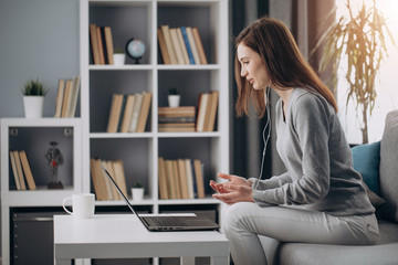 Side view of beautiful girl in casual clothing sitting on couch and having video chat. Charming woman using laptop and headphones for online conversation at home.