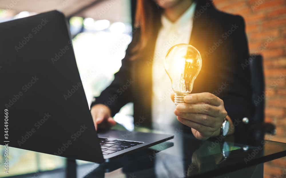 Wall mural closeup image of a businesswoman holding a glowing light bulb while working on laptop computer in of