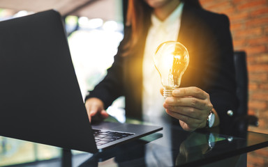 Closeup image of a businesswoman holding a glowing light bulb while working on laptop computer in...